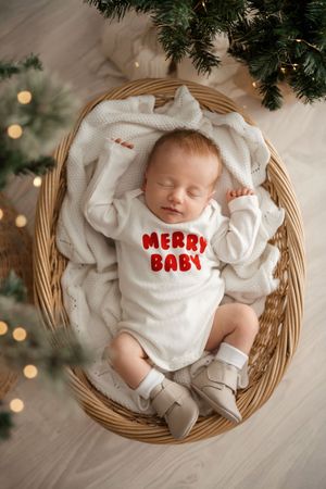 A baby wearing a white outfit with 'Merry Baby' text lies in a round wicker basket under Christmas tree branches.