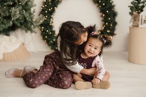 Two children share a sweet embrace while sitting on the floor with Christmas decorations in the background.