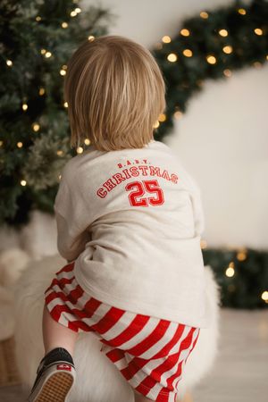 Child wearing Christmas pajamas with number 25 sits near a decorated tree with twinkling lights in the background.