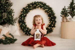 A holiday photo session featuring a small child in a red dress sitting on a white floor with Christmas wreaths in background.