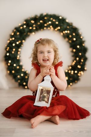 Festive holiday portrait with red dress and white lantern in front of illuminated Christmas wreath.