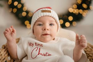Baby wearing white knit hat and 'My First Christmas' outfit lying down with holiday lights bokeh in background.