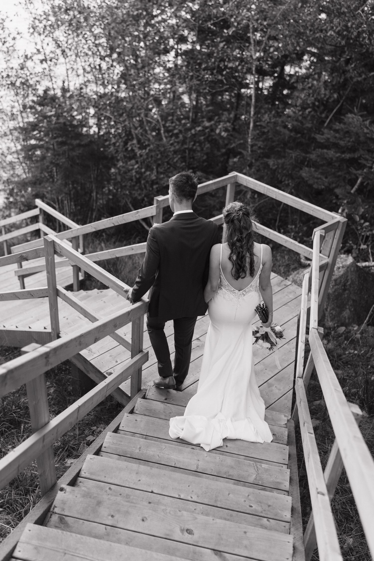 Bride and groom walking up wooden stairs surrounded by forest in black and white at private family estate in Nova Scotia, Canada.