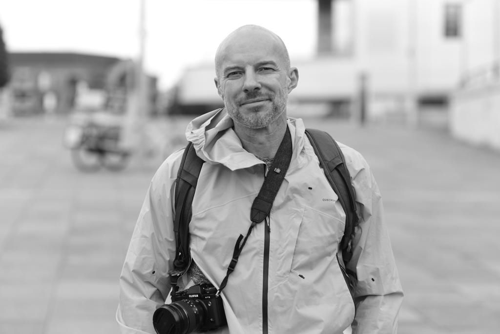 Black and white portrait of Photographer Gary Williams with camera and backpack outdoors.