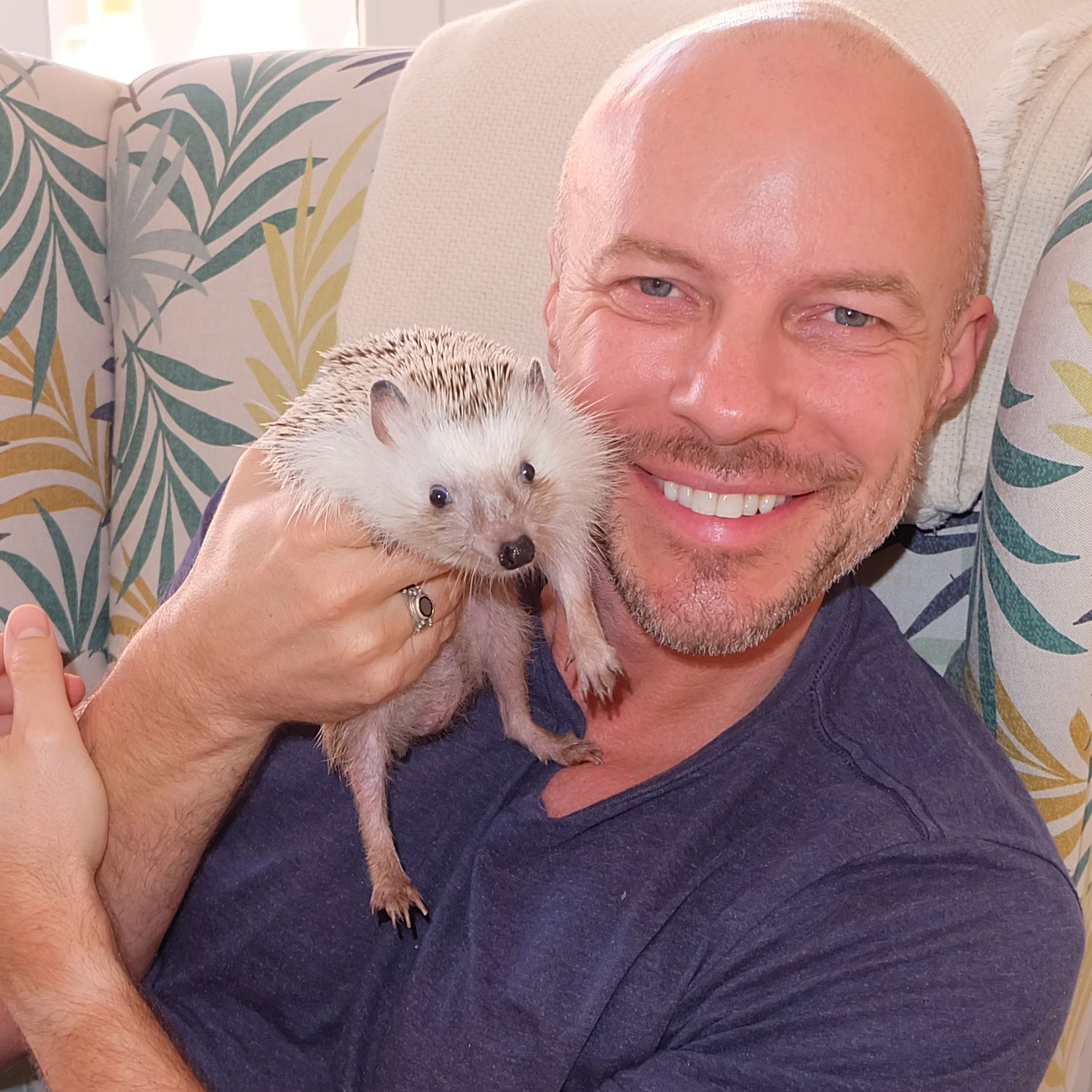 Photographer Gary Williams holding small white hedgehog pet against floral patterned background.