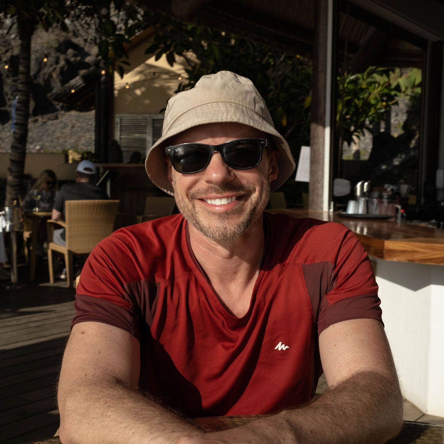 Photographer Gary Williams wearing a burgundy v-neck t-shirt, bucket hat and sunglasses smiles while sitting outdoors at a cafe.