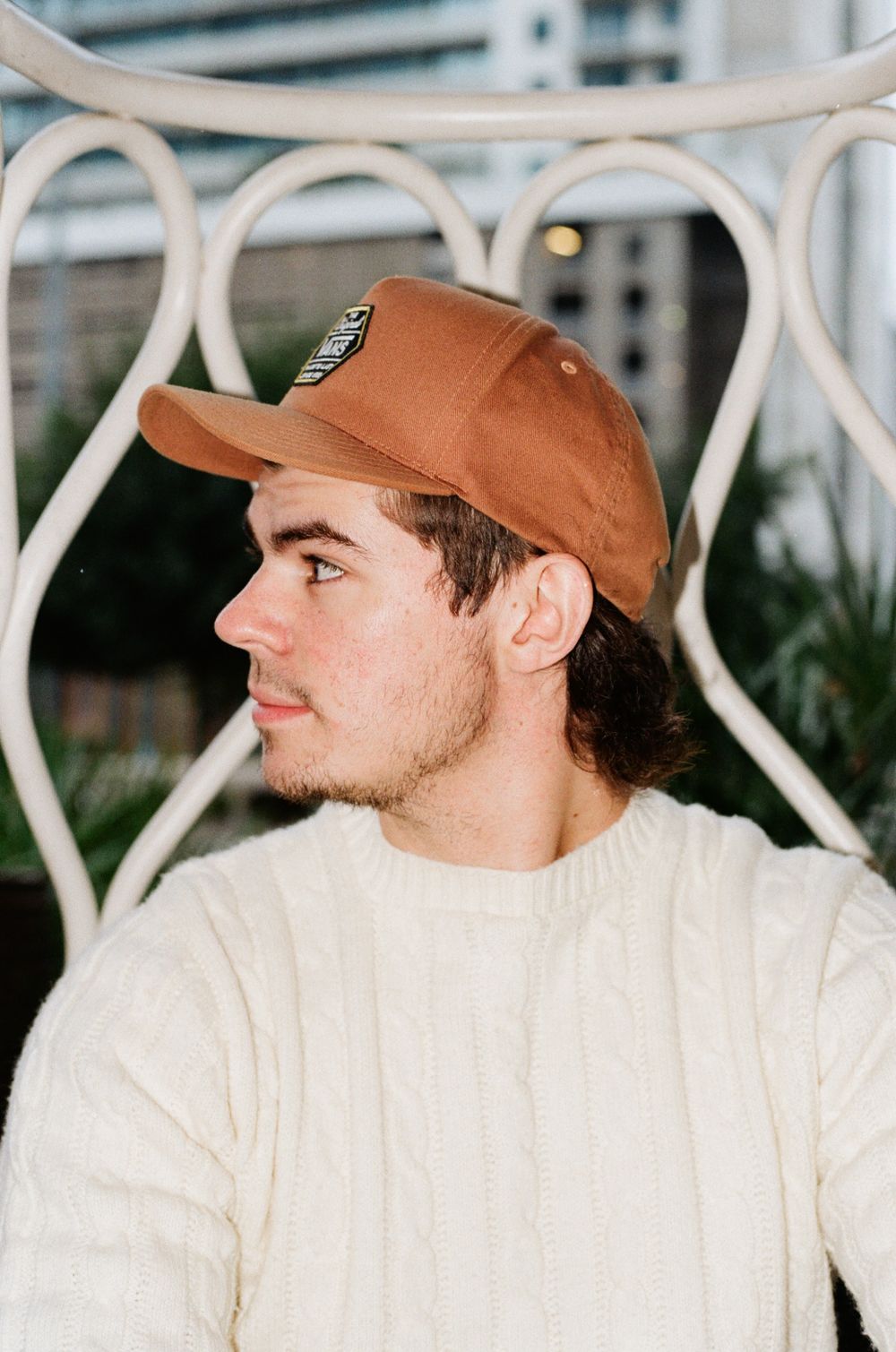 Person in cream sweater and orange baseball cap poses in profile against white metal patio furniture.