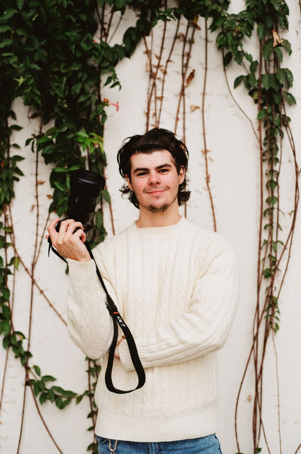 Person in cream sweater holding camera while standing against ivy-covered wall.