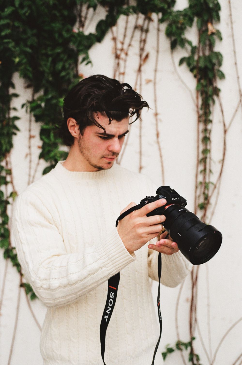 Person in white cable knit sweater examining camera settings against vine-covered wall.