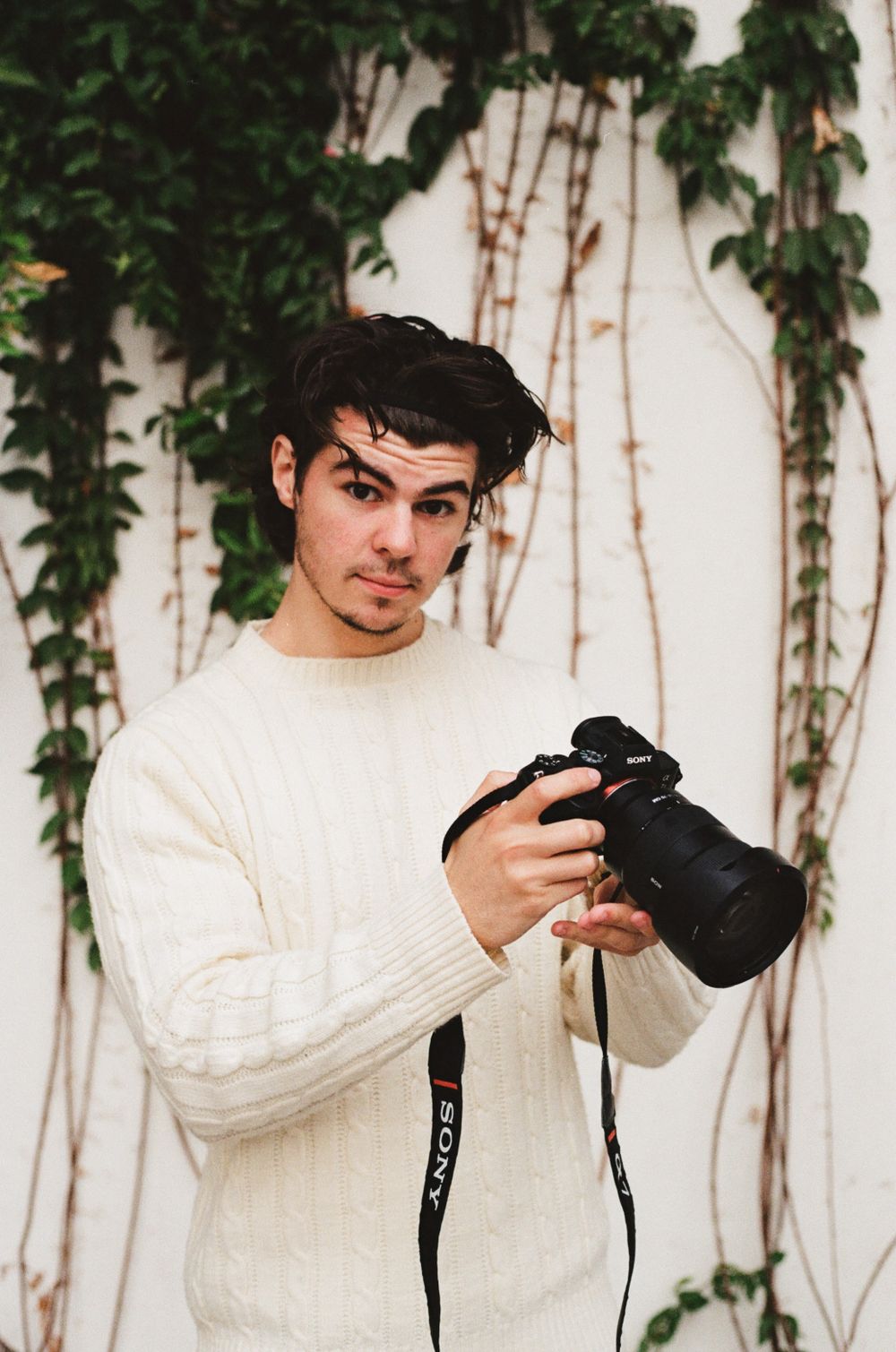 Person holding professional camera while wearing white sweater against wall with climbing vines.