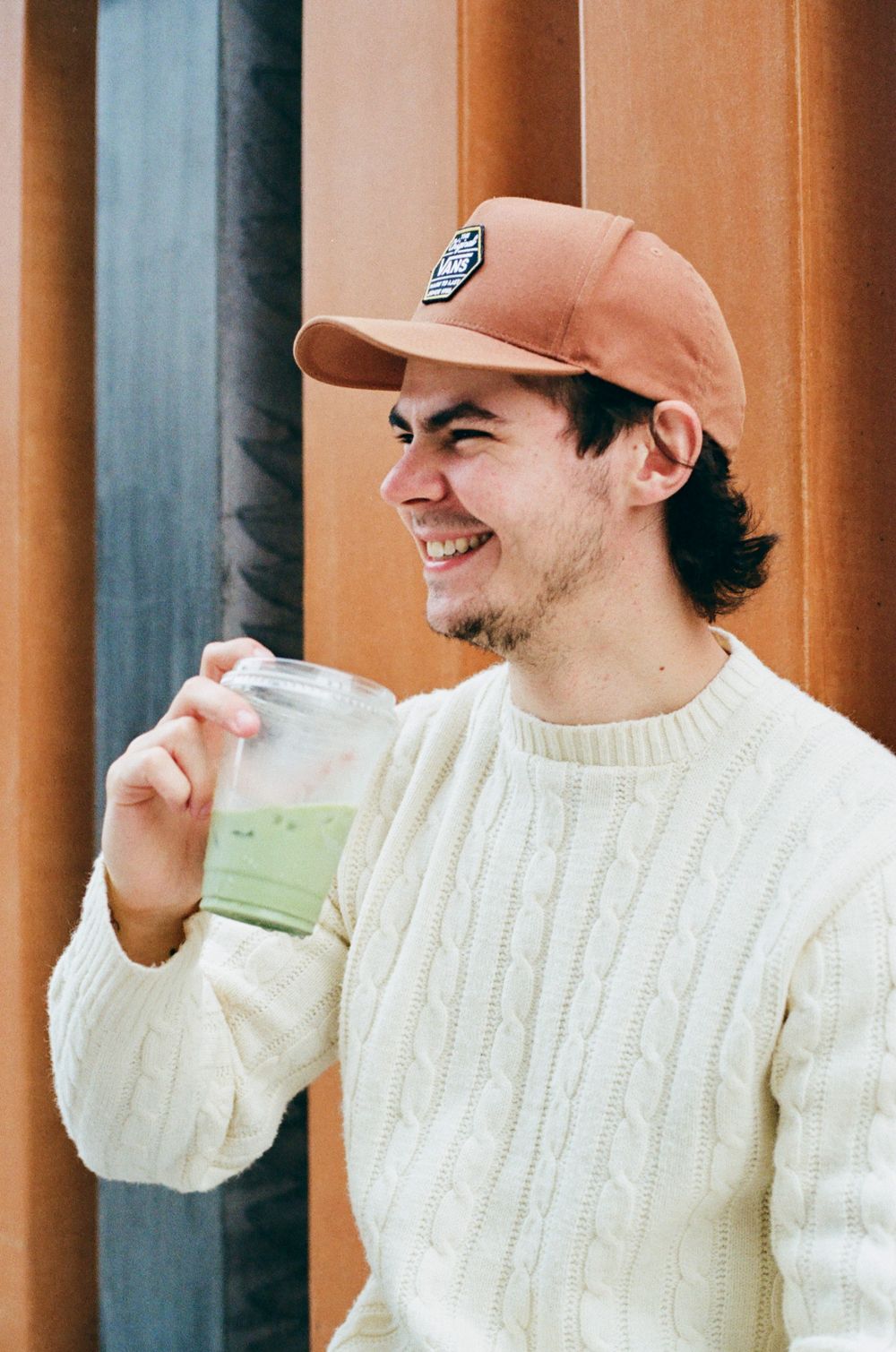 Person in white sweater and tan cap smiling while holding matcha outdoors.