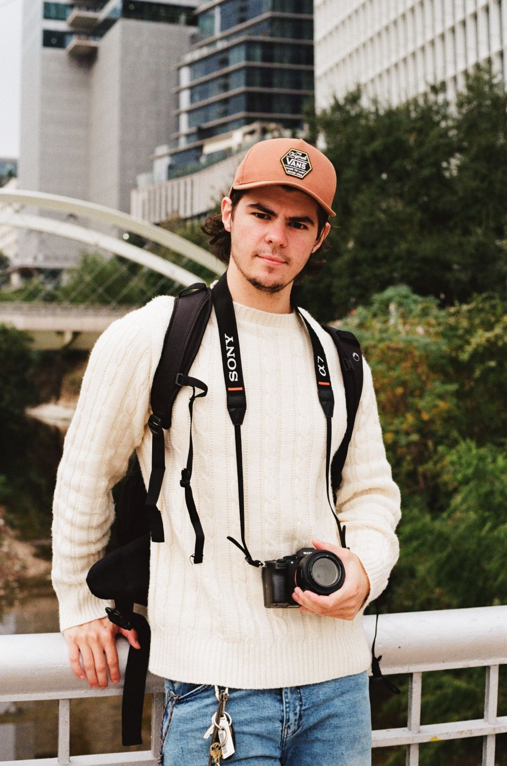 Photographer in white sweater and orange cap with camera gear against urban backdrop downtown Austin.