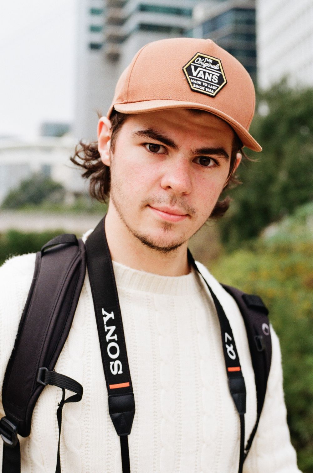 Photographer wearing Vans cap and camera strap poses with photography equipment outdoors.