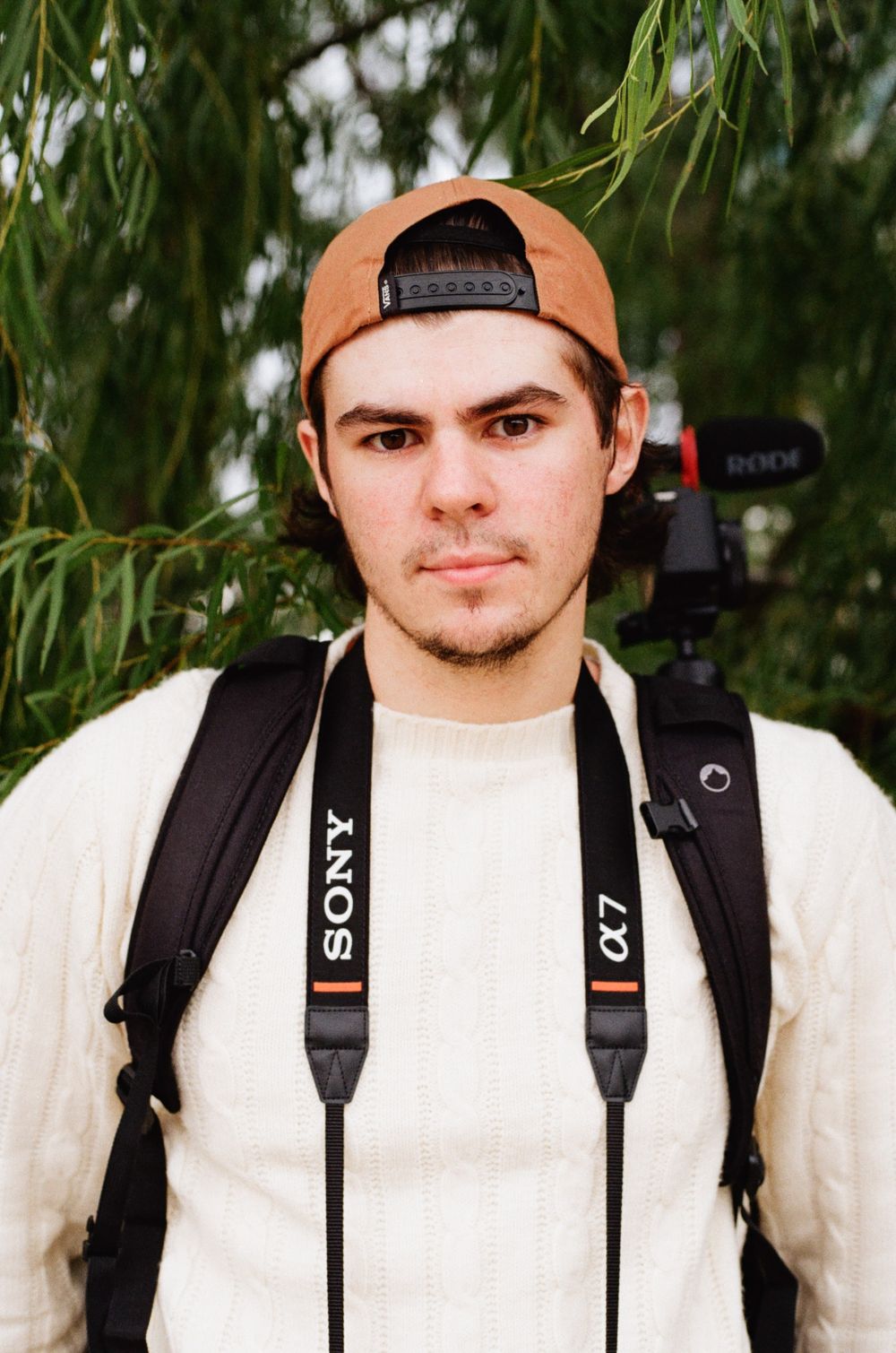 Photographer with Sony camera straps wearing a backwards cap and white sweater against greenery.