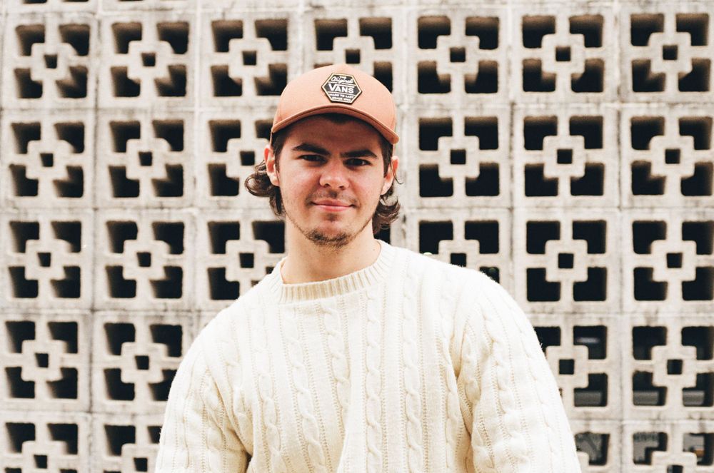Photographer in cream cable knit sweater and baseball cap against decorative concrete wall.