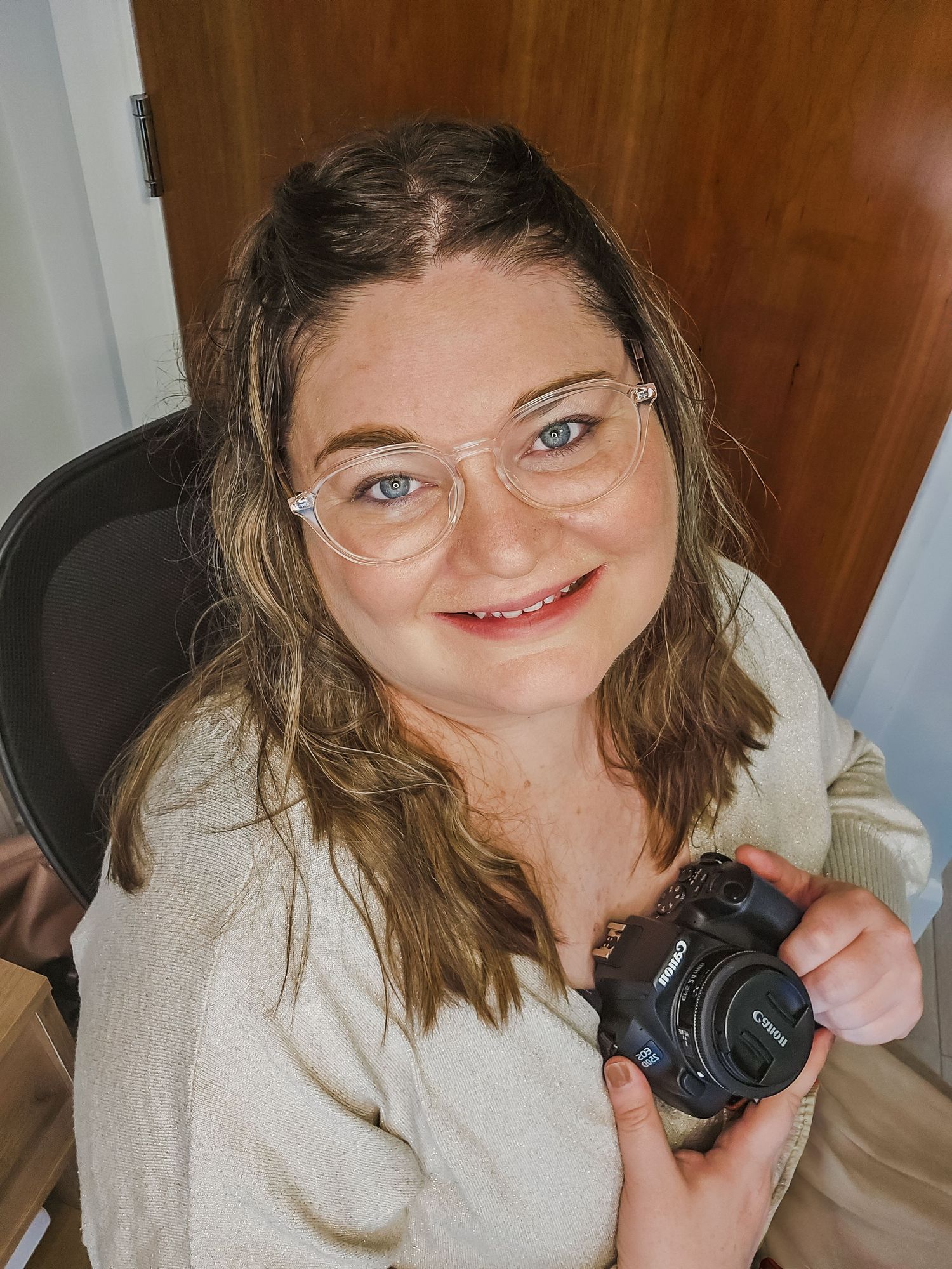 A photographer wearing a cream sweater and glasses holds up a camera while seated in a black office chair.