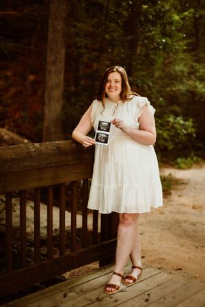 Woman in white dress stands on wooden bridge holding ultrasound photos.