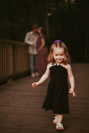 Little girl in a black dress dances playfully on a wooden walkway at sunset.