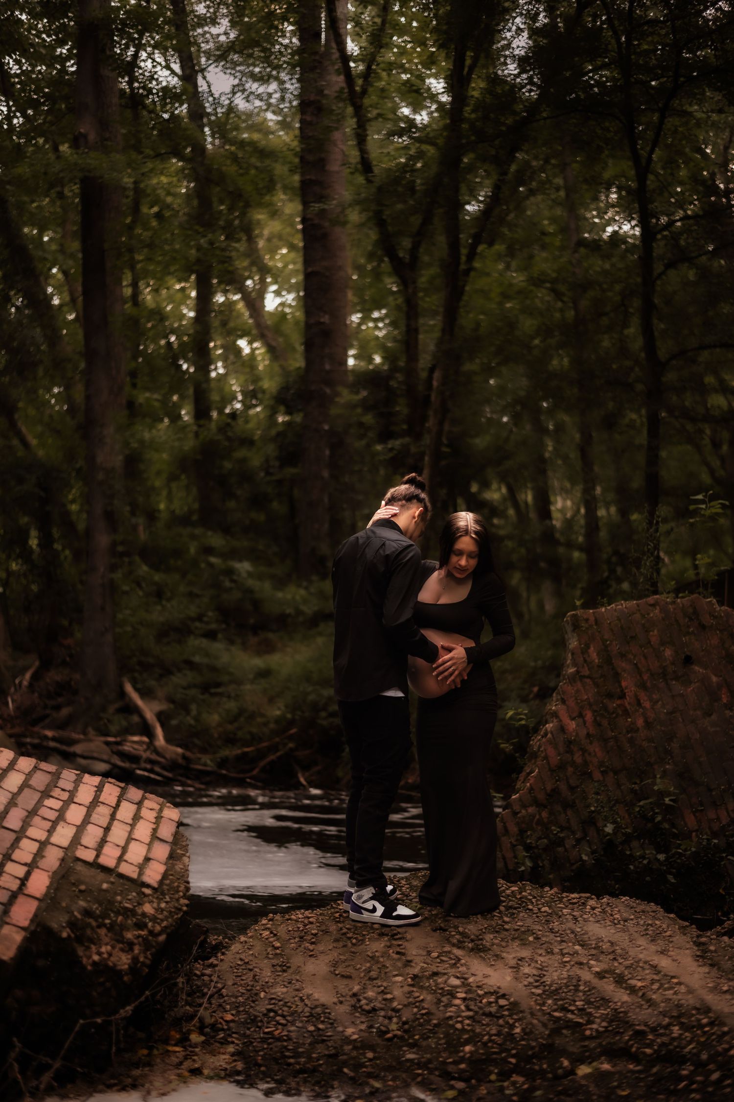Couple embracing on dirt path in moody forest setting.