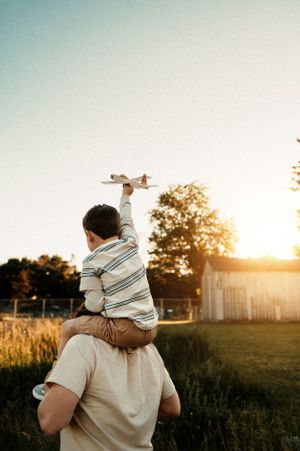 Child riding on shoulders while holding toy airplane at sunset in backyard.