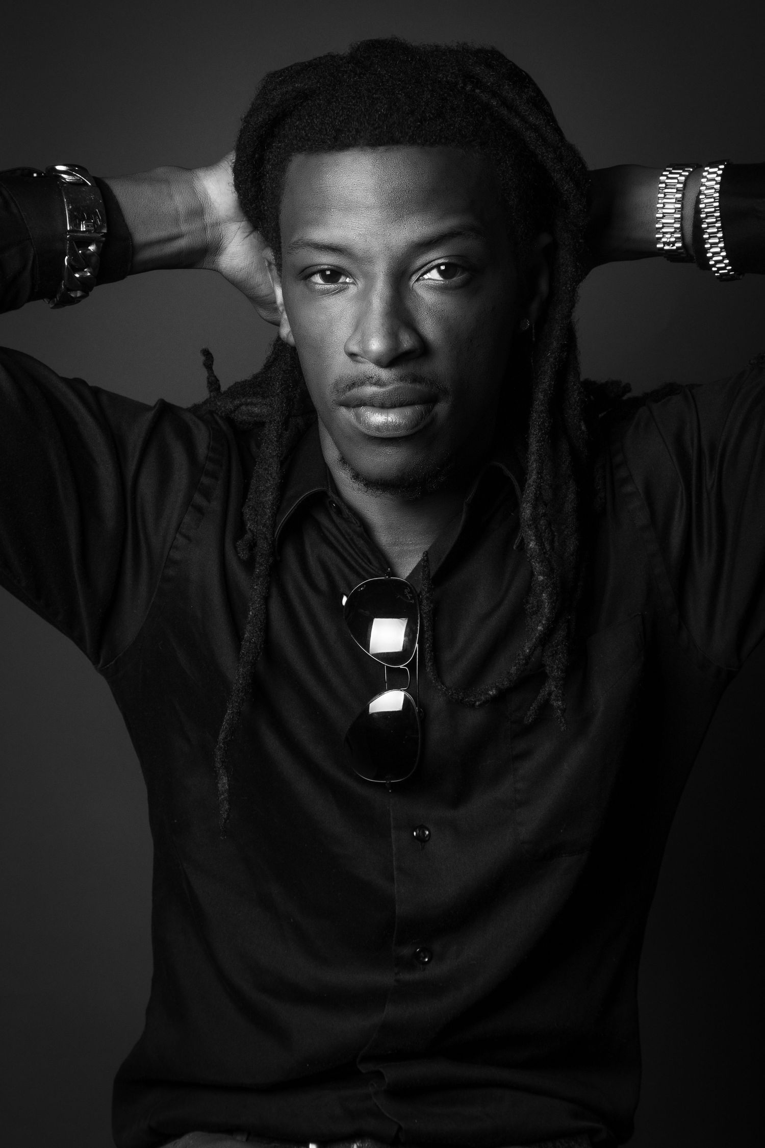 A black and white portrait of someone in a dark shirt with hands raised behind their head against a dark background.