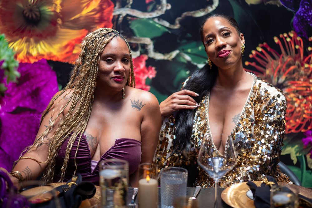 Two friends enjoying drinks at a colorful party with floral decorations.