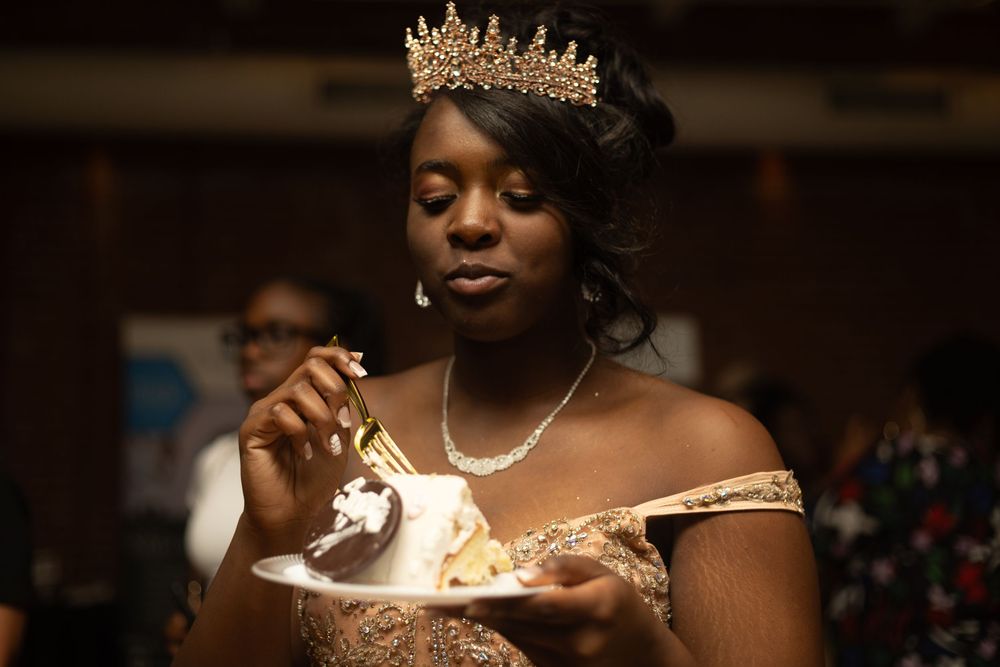 An elegant figure in a gold dress and sparkling tiara enjoys a slice of cake at a formal celebration.