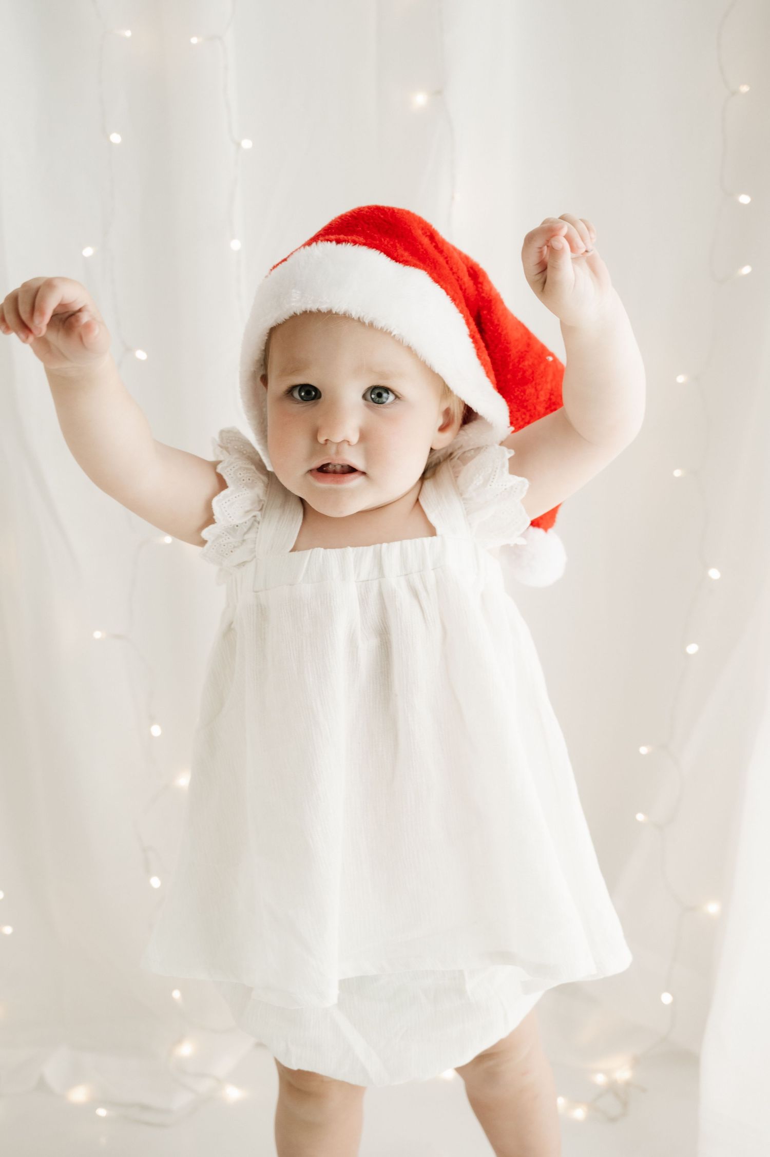 Baby in white dress and Santa hat reaching up against sparkly backdrop.