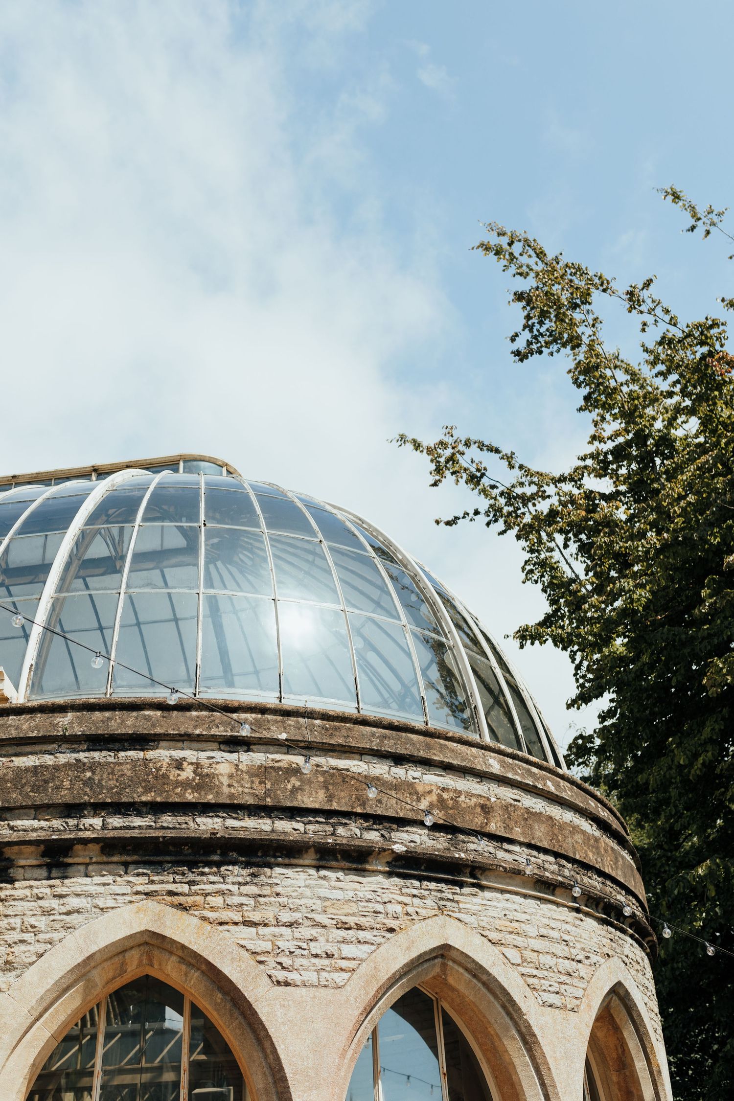 Glass dome atop a historic stone building with gothic arched windows against a bright blue sky with tree branches.