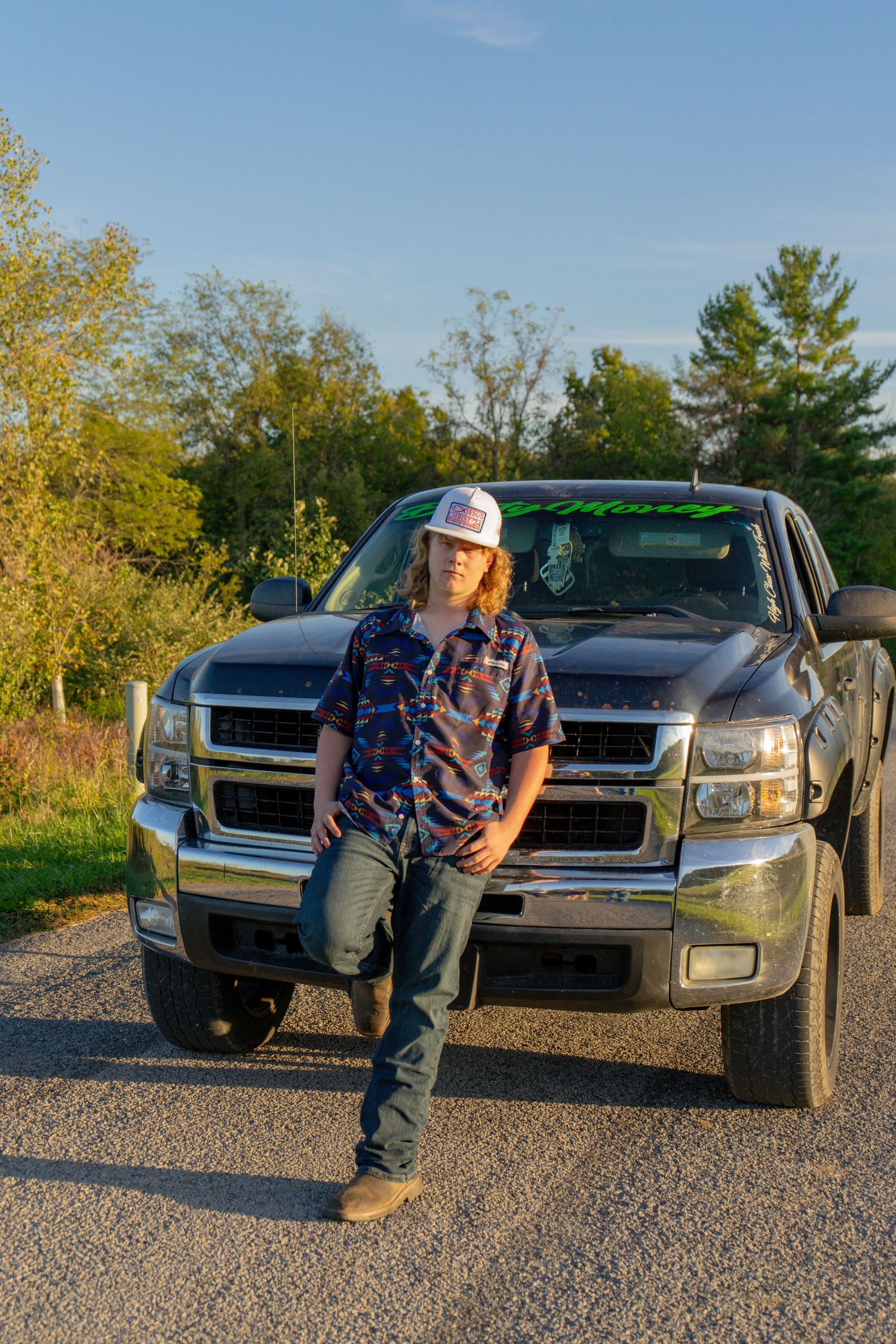 Young man in flannel shirt and trucker hat leaning against a black pickup truck on a gravel road.