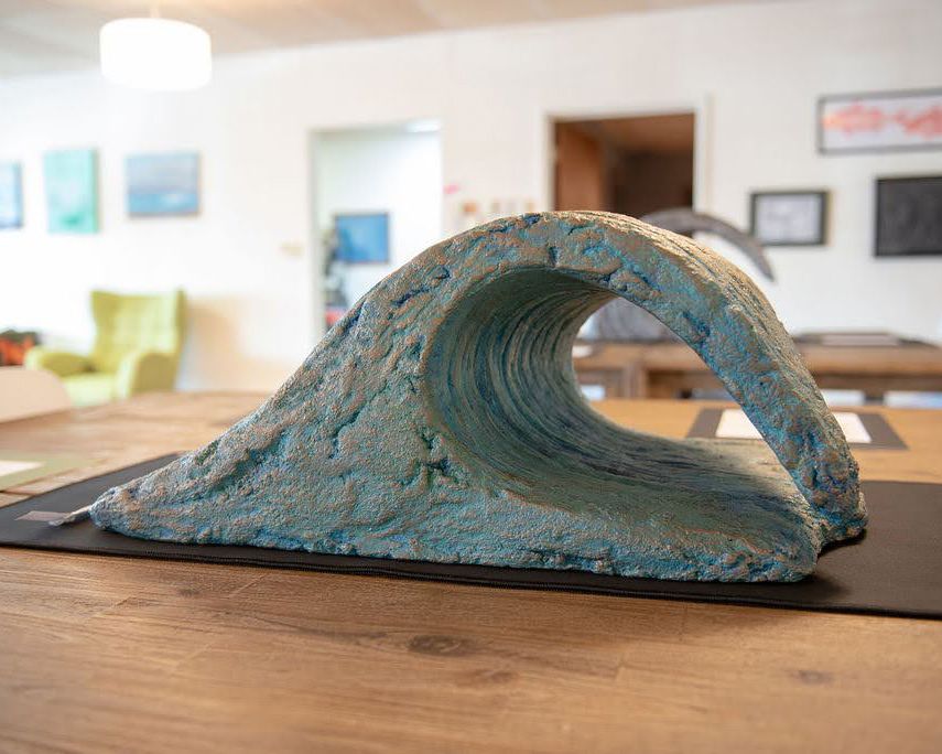 A sculptural art piece depicting a curling ocean wave made from blue-tinted material on a wooden surface.