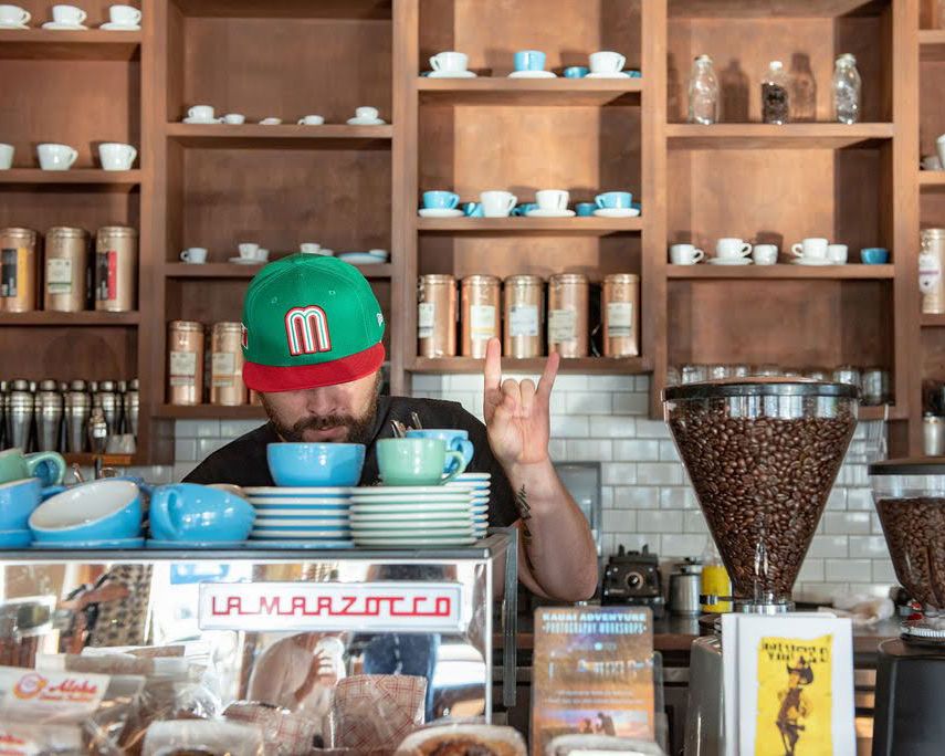 A barista in a green cap serves coffee at a rustic cafe counter with wooden shelves filled with coffee supplies.