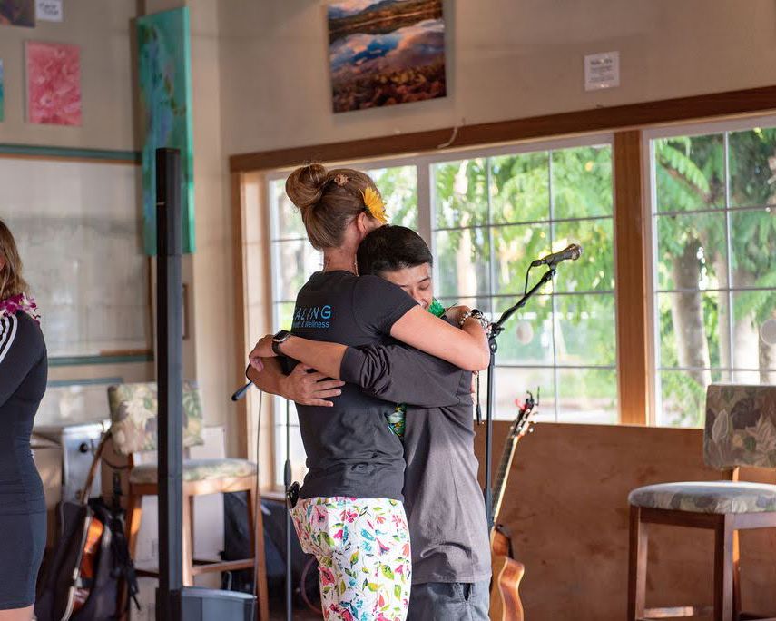 An emotional embrace between performers in a sunlit venue with large windows.