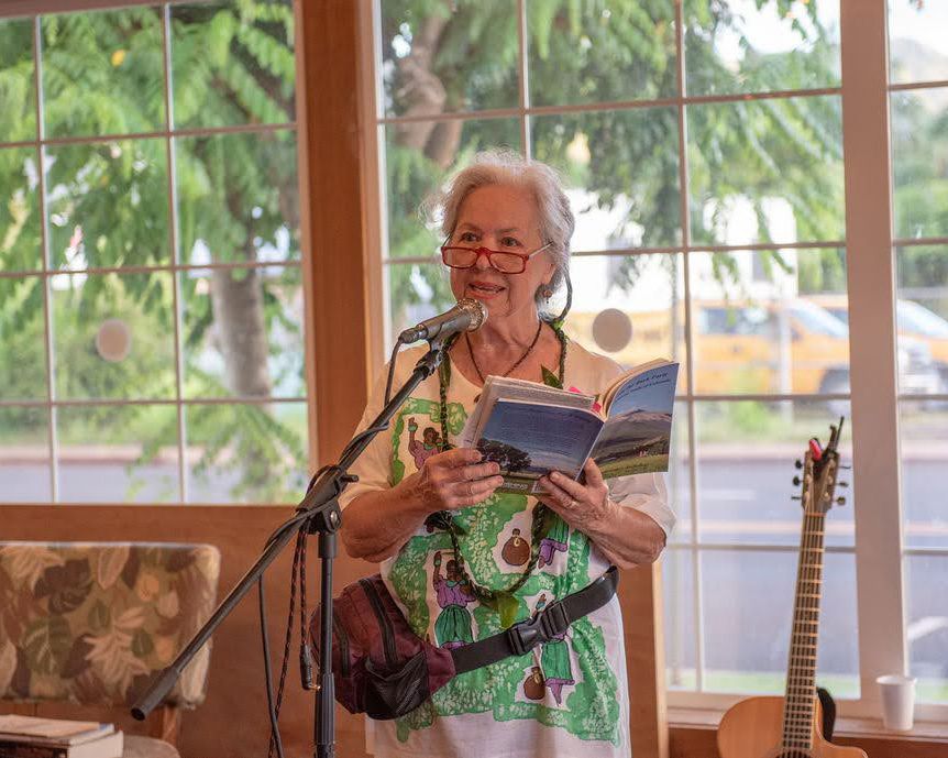 A performer in tropical attire reads from a book while standing at a microphone by sunny windows.