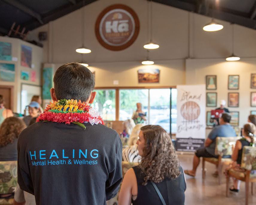 A performer wearing a HEALING shirt stands in front of a cafe logo and scenic artwork.