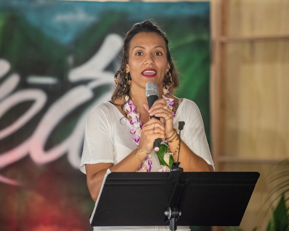 Speaker at podium holds microphone during performance against tropical backdrop.