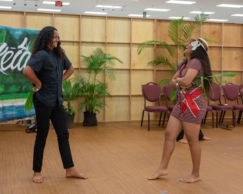 Two performers practicing a dance routine in a room with tropical plants and chairs.
