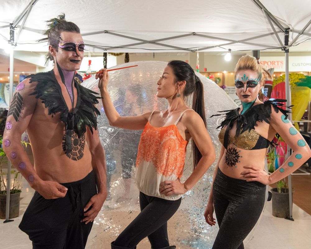 Three performers in colorful festival attire laugh and dance together at an outdoor market event.