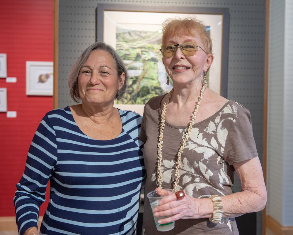 Two friends pose together at an art gallery event wearing casual attire.