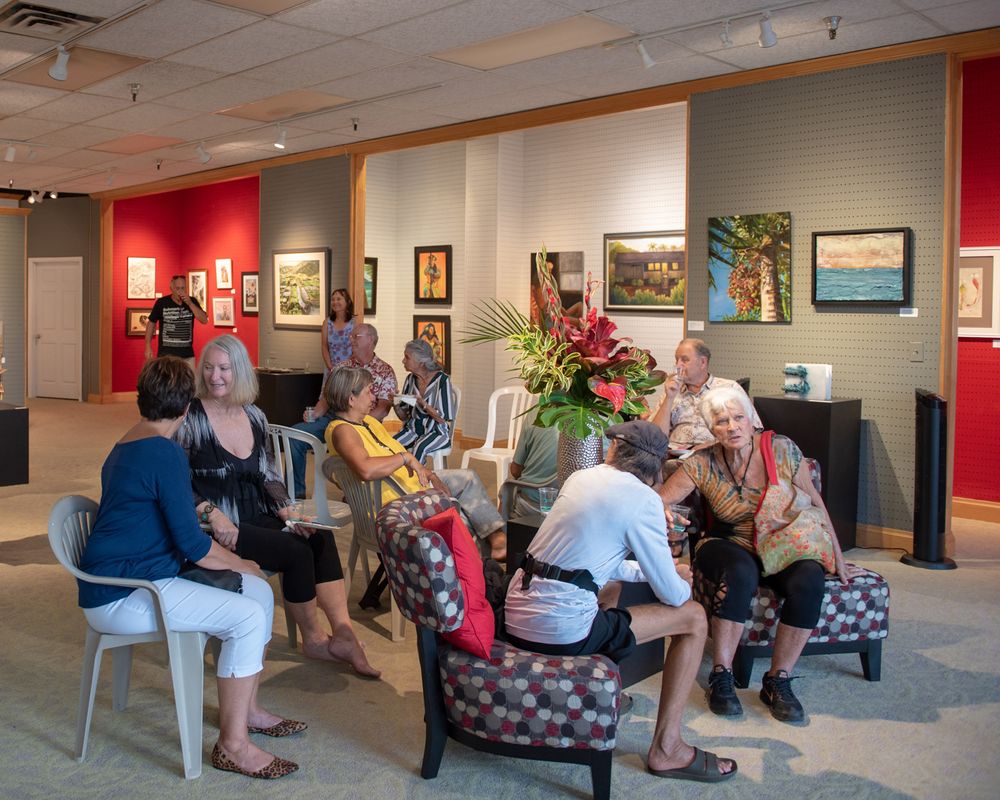 A group gathers in chairs at an art gallery surrounded by paintings on display.