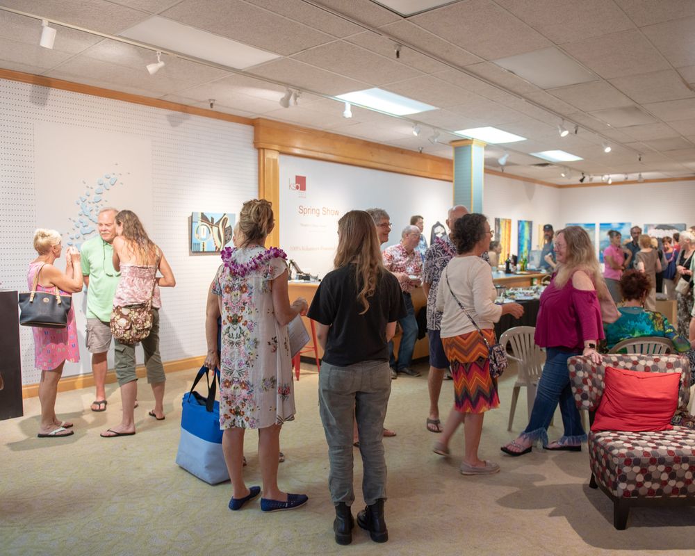 Crowd of visitors browse art and merchandise displays at indoor gallery event.