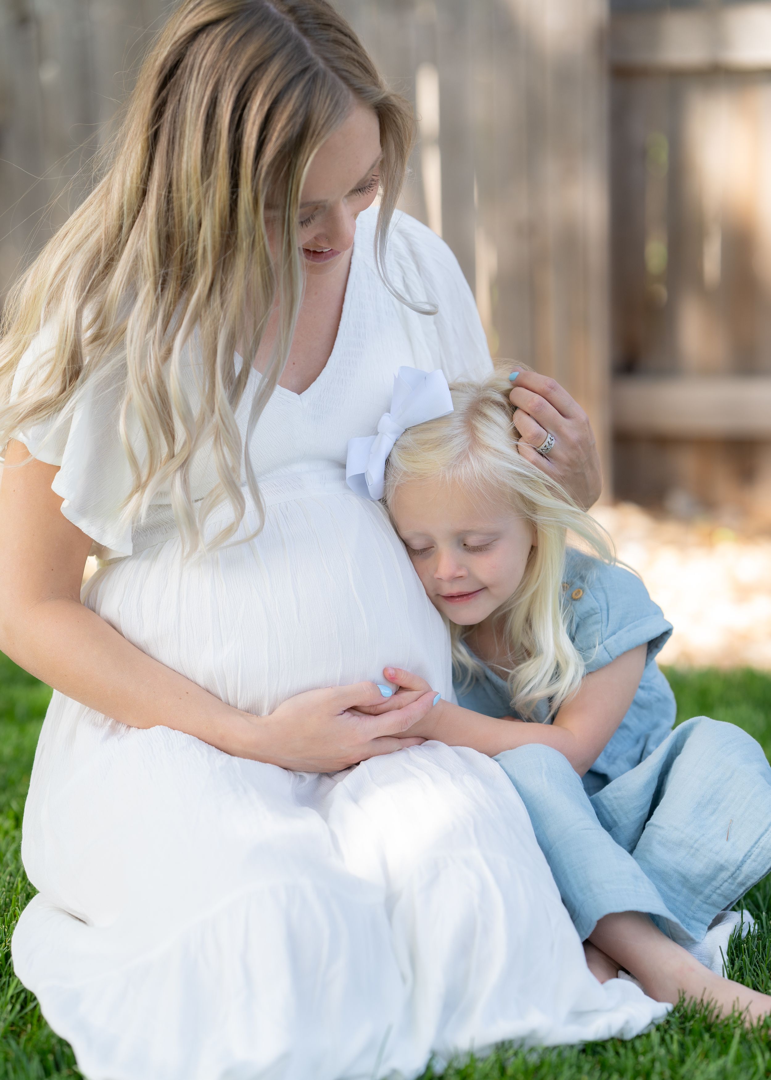 Maternity photo of pregnant mother in white dress sitting on grass with young child.