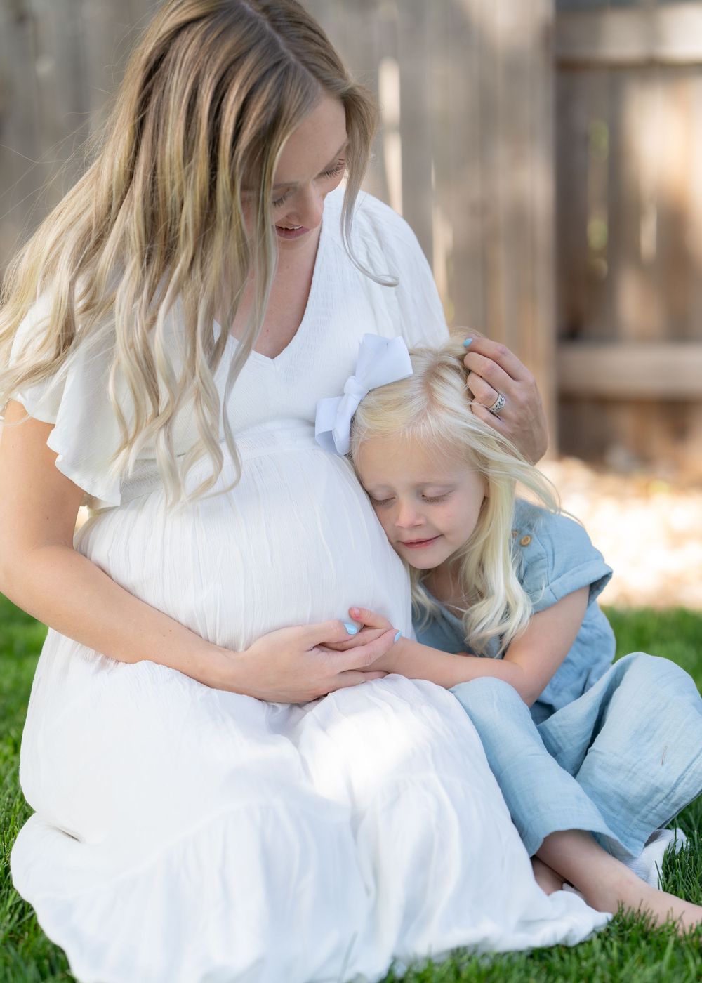Maternity photo of pregnant mother in white dress sitting on grass with young child.