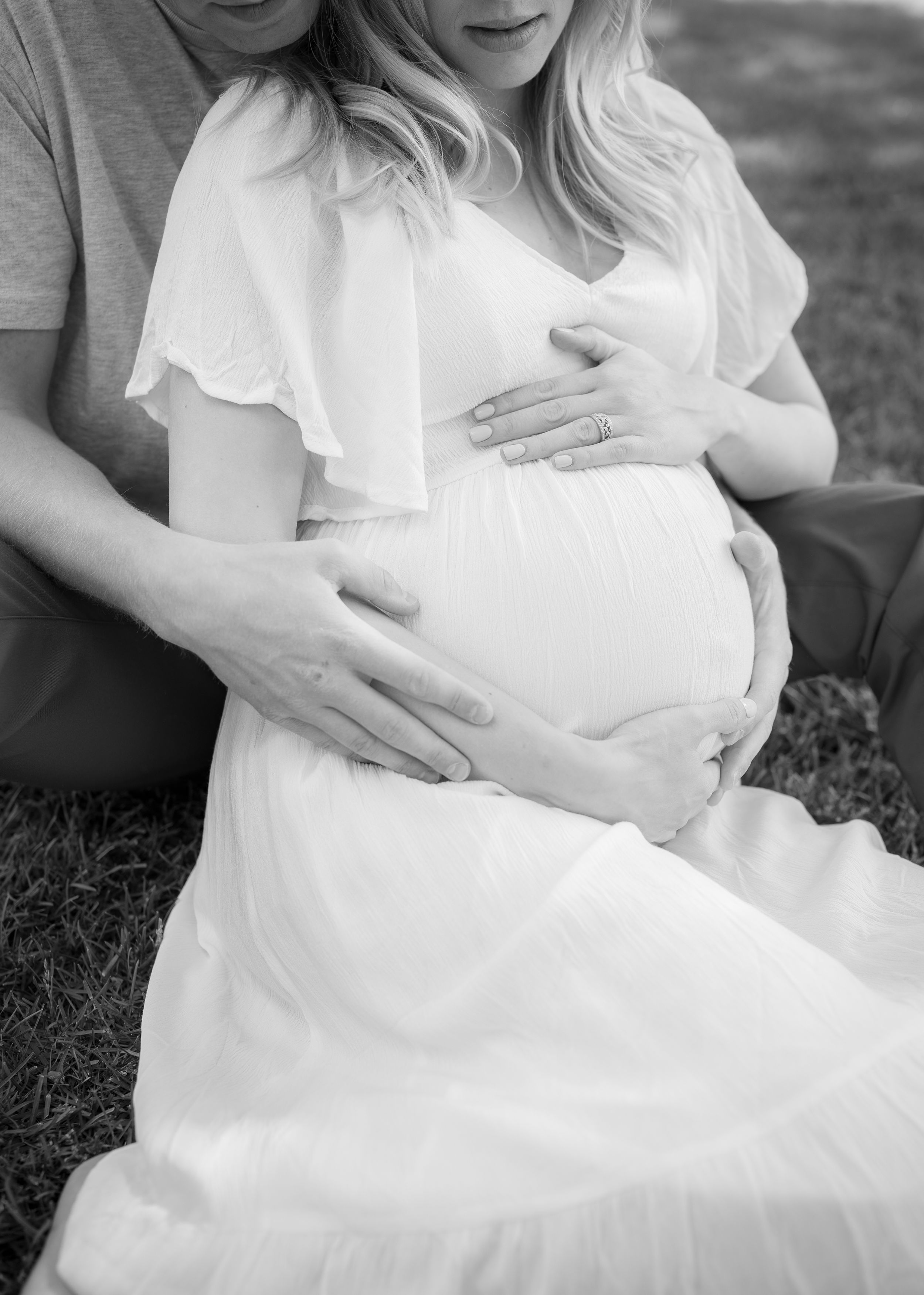 Black and white photo of young couple cradling pregnant belly in flowing white dress.