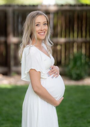 Maternity portrait of young woman in white dress against wooden fence in garden setting.