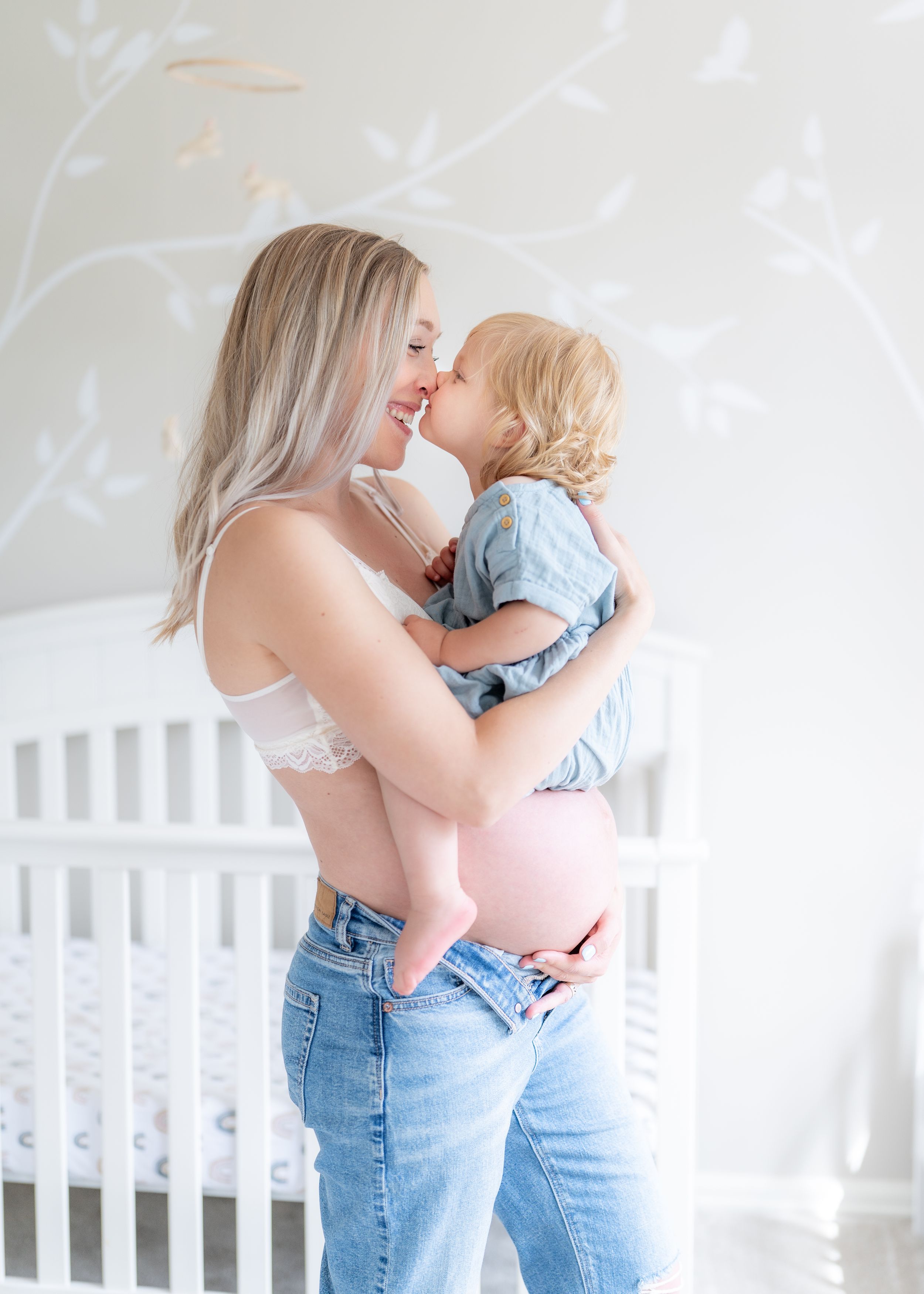 Maternity photo of pregnant mother and child sharing affectionate moment in front of white crib.
