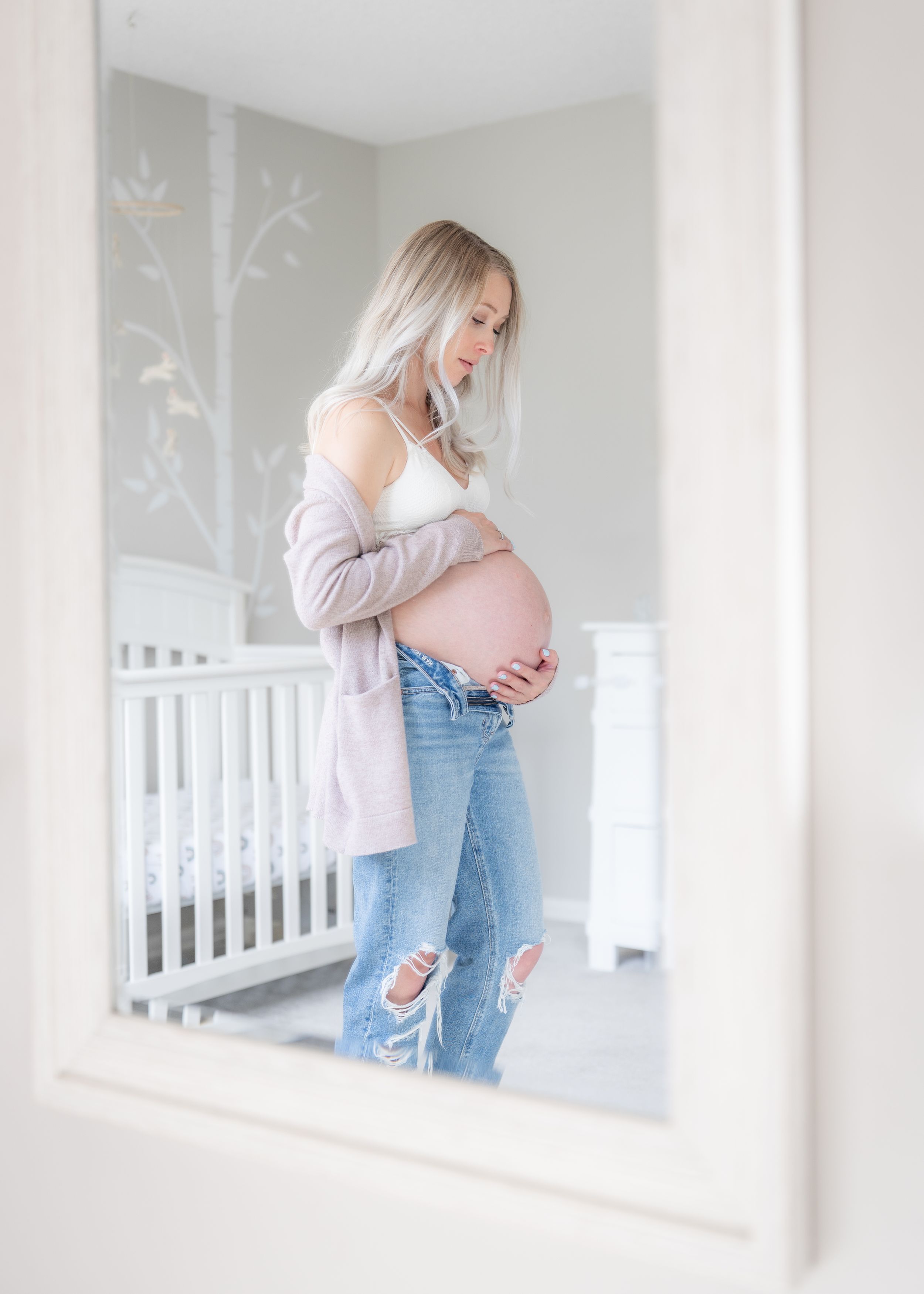 Maternity photo of an expectant mother in ripped jeans and beige cardigan standing in a nursery with white crib.