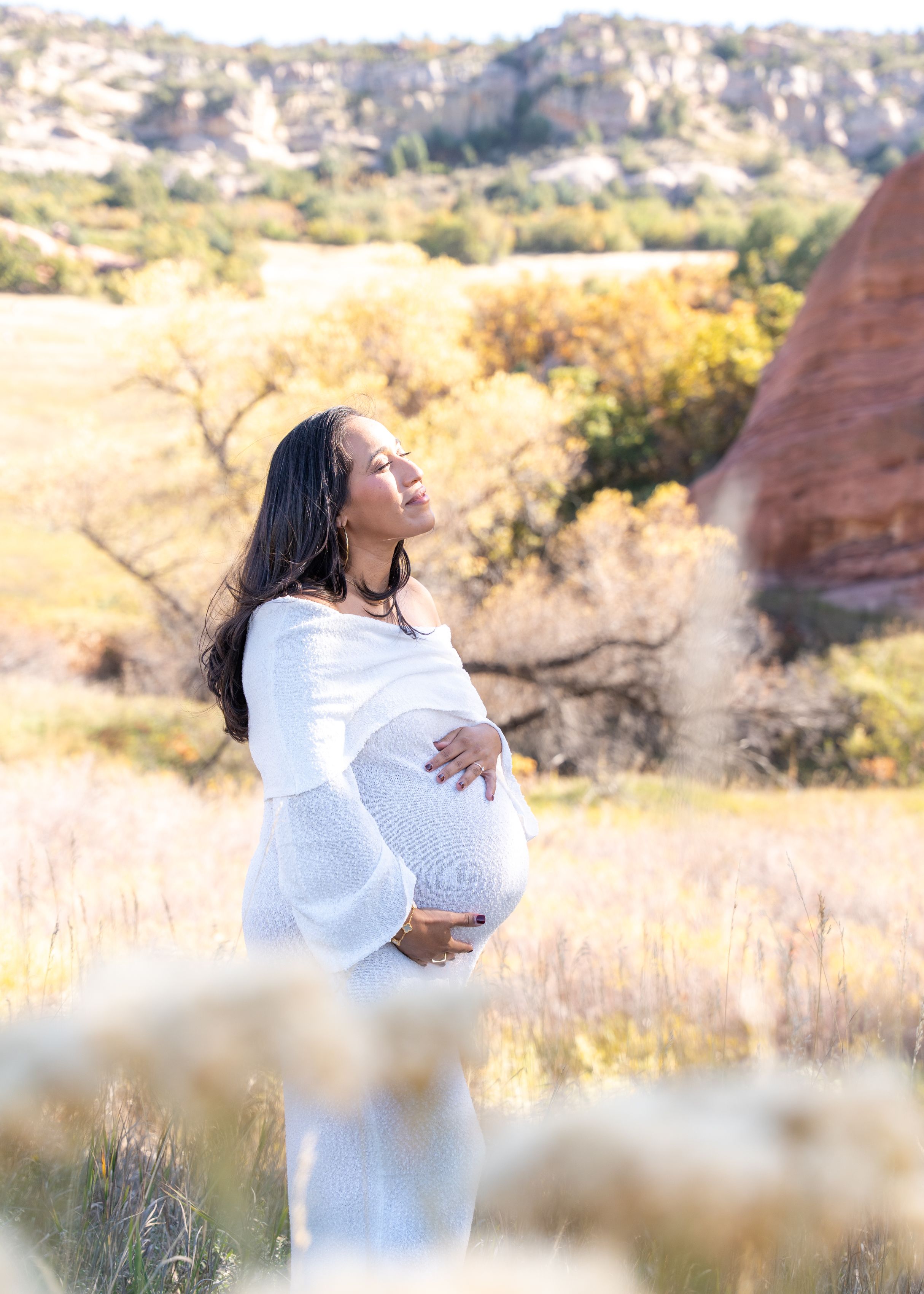 Pregnant woman in white dress poses outdoors in golden sunlight during maternity photoshoot.