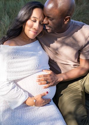 Couple embraces during outdoor maternity photoshoot with woman wearing white dress.