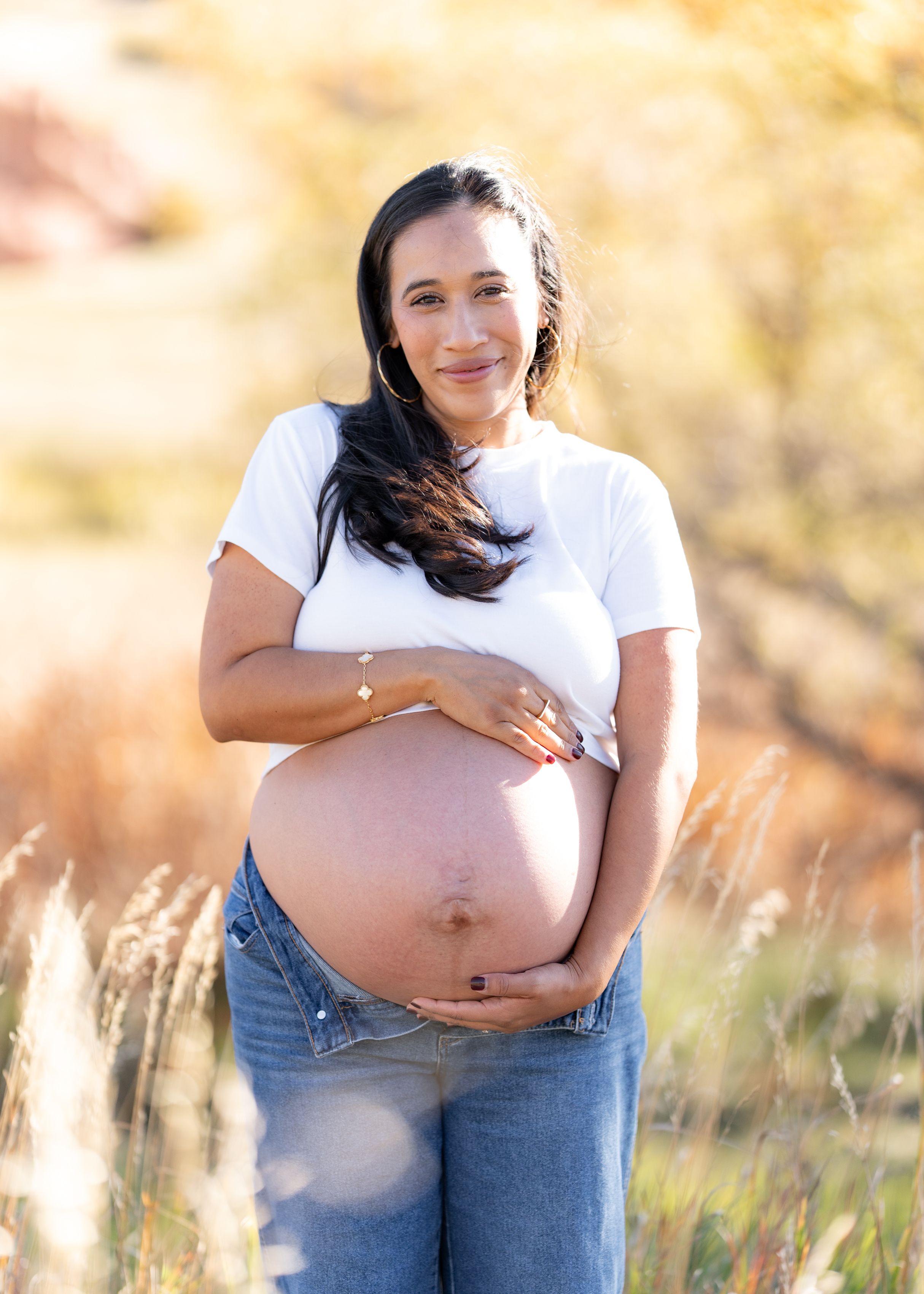 A pregnant woman in a white t-shirt and jeans poses in a sunny field during golden hour.